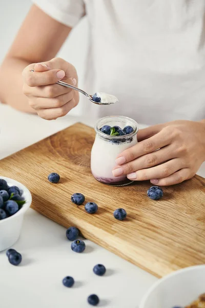 Cropped shot of woman at tabletop with yogurt with fresh blueberries for breakfast on wooden cutting board — Stock Photo
