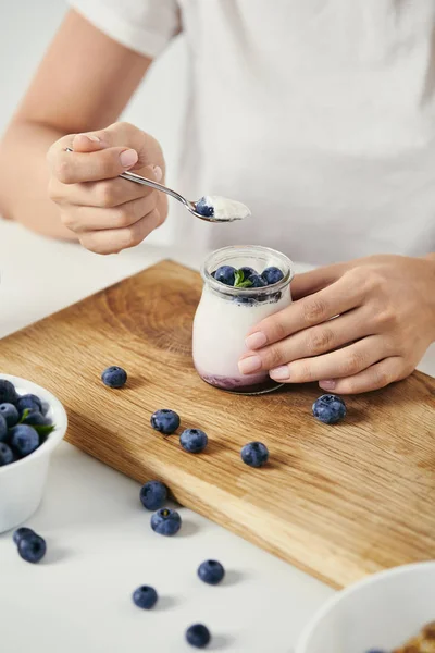 Abgeschnittene Aufnahme einer Frau an der Tischplatte mit Joghurt mit frischen Blaubeeren zum Frühstück auf einem Holzschneidebrett — Stockfoto