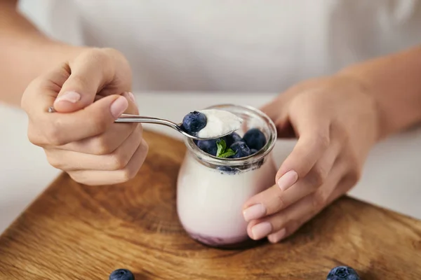 Colpo ritagliato di donna al tavolo con yogurt con mirtilli freschi per la prima colazione sul tagliere di legno — Foto stock