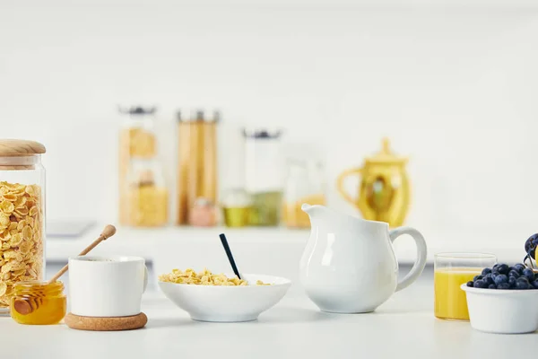 Vista ravvicinata di fiocchi di mais in ciotola, tazza di caffè e brocca di latte per la colazione sulla superficie bianca — Foto stock