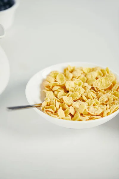 Cerrar la vista de cuchara y copos de maíz con leche en tazón para el desayuno en la superficie blanca - foto de stock