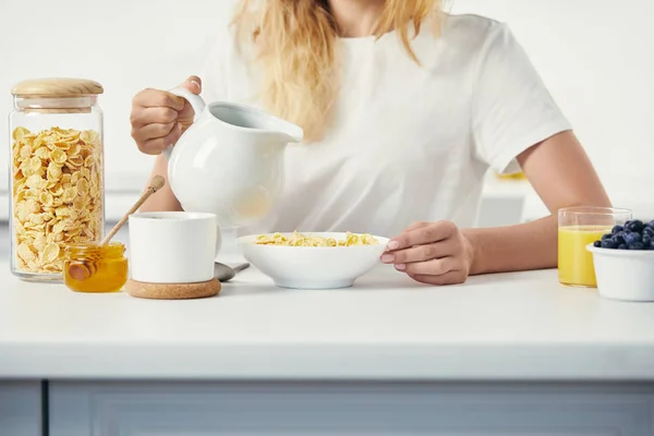 Teilansicht einer Frau mit Milchkrug am Tisch sitzend mit einer Schüssel Cornflakes und einem Glas Saft zum Frühstück zu Hause — Stockfoto