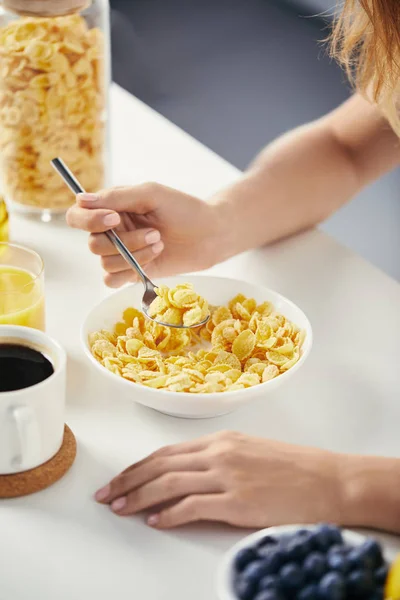 Partial view of woman with spoon sitting at table with corn flakes and cup of coffee for breakfast — Stock Photo