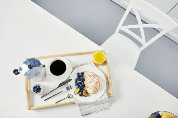 Vista dall'alto di croissant con mirtilli freschi e pezzi di prugne, bicchiere di succo di frutta, tazza di caffè e yogurt su vassoio di legno per colazione su tavolo bianco — Foto stock