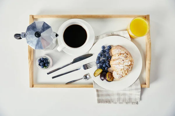 Draufsicht auf Croissant mit frischen Blaubeeren und Pflaumenstücken, Glas Saft, Tasse Kaffee und Joghurt auf Holztablett zum Frühstück auf weißer Tischplatte — Stockfoto