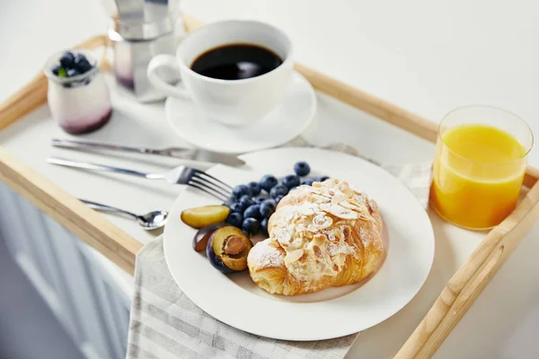 Vista de perto de croissant com mirtilos frescos e pedaços de ameixa, copo de suco, xícara de café e iogurte para café da manhã em bandeja de madeira em mesa branca — Fotografia de Stock