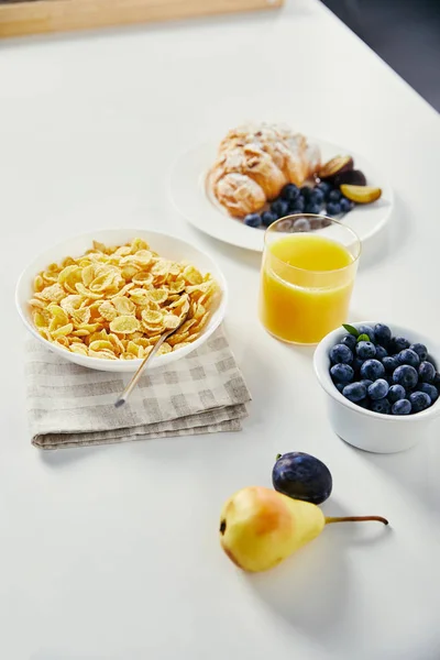 Close up view of corn flakes in bowl, glass of juice and croissant with blueberries and plum pieces for breakfast on white tabletop — Stock Photo