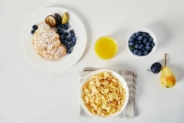 Plat déposer avec des flocons de maïs dans un bol, verre de jus et croissant avec des bleuets et des morceaux de prune pour le petit déjeuner sur la table blanche — Photo de stock