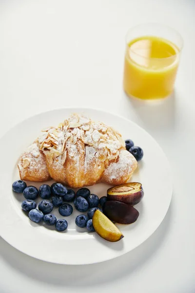 Close up view of glass of juice and croissant with blueberries and plum pieces for breakfast on white tabletop — Stock Photo