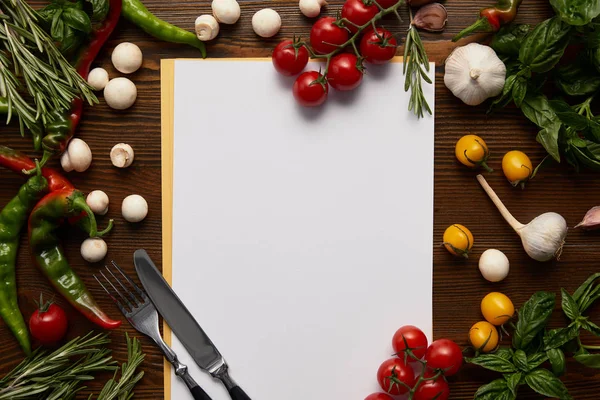 Vista superior de la tarjeta en blanco, cubiertos y hierbas frescas con verduras en la superficie de madera - foto de stock