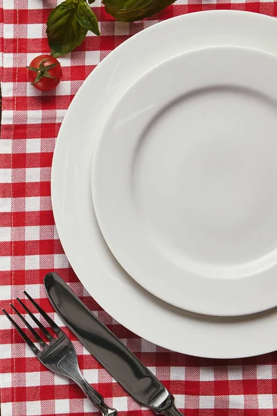 Top view of empty white plates, cutlery and fresh basil with tomato on checkered tablecloth — Stock Photo