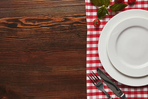 Top view of empty white plates, cutlery and fresh basil with tomatoes on wooden surface — Stock Photo