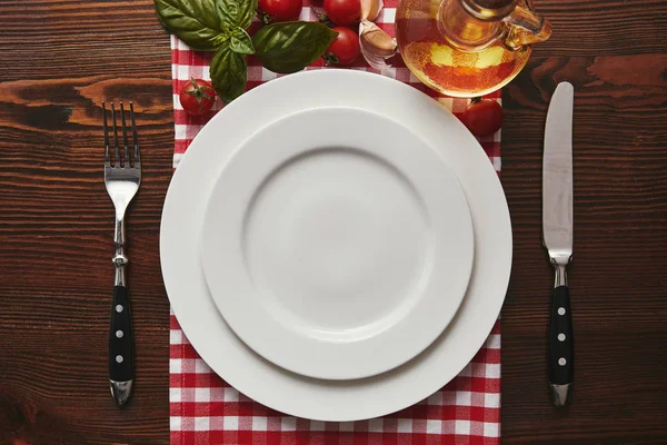 Top view of empty white plates, cutlery and fresh basil with tomatoes and oil on wooden surface — Stock Photo