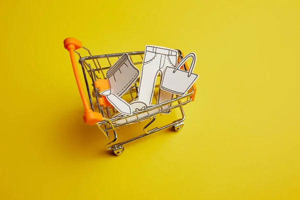 Vista de cerca del pequeño carrito de compras con ropa hecha de papel sobre fondo amarillo - foto de stock