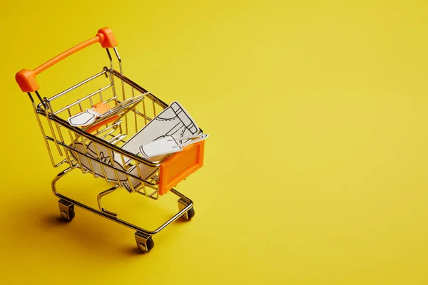 Vista de cerca del pequeño carrito de compras con ropa hecha de papel sobre fondo amarillo - foto de stock