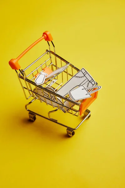 Vista de cerca del pequeño carrito de compras con ropa hecha de papel sobre fondo amarillo - foto de stock