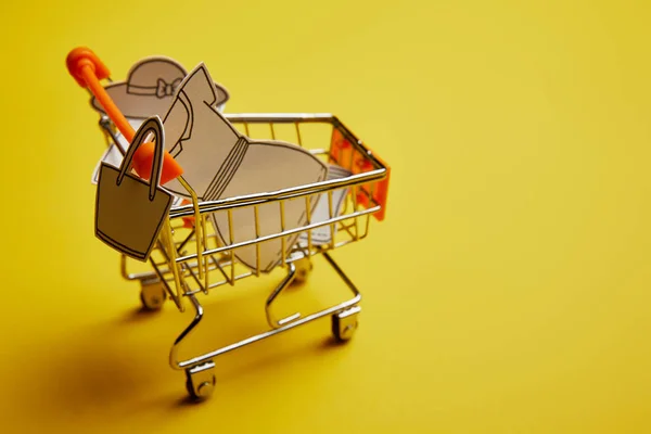 Close up view of little shopping trolley with paper clothes on yellow backdrop — Stock Photo