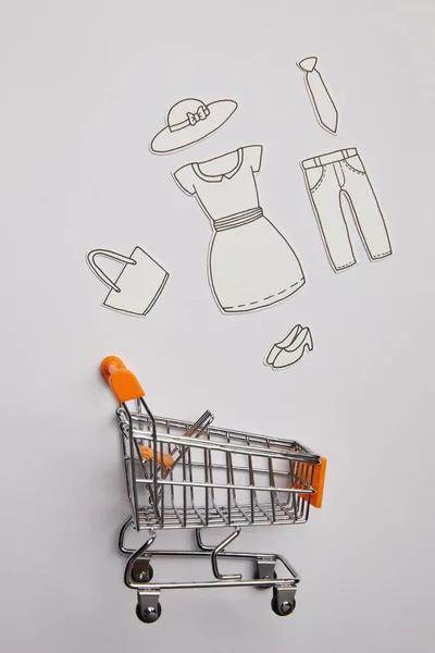 Top view of shopping cart and little clothes made of paper on grey background — Stock Photo