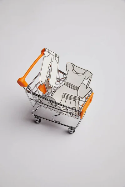 Close up view of shopping cart with little clothes made of paper on grey background — Stock Photo