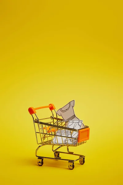 Vista de cerca del pequeño carrito de compras con ropa de papel sobre fondo amarillo - foto de stock