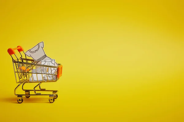 Close up view of little shopping trolley with paper clothes on yellow backdrop — Stock Photo