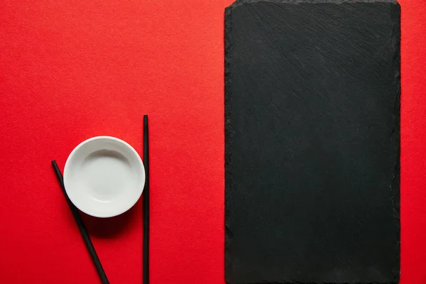 Top view of arranged empty bowl, chopsticks and slate plate on red background — Stock Photo