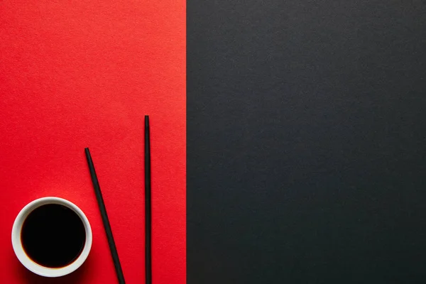 Vista superior de molho de soja na tigela e pauzinhos no fundo vermelho e preto — Fotografia de Stock