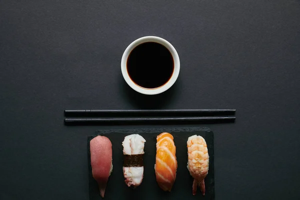 Вид сверху на набор суши из нигири, палочки для еды и соевый соус на тарелке из черного шифера на черном столе — стоковое фото