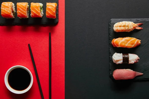 Acostado plano con salsa de soja en un tazón, palillos y conjuntos de sushi en platos de pizarra negra sobre fondo rojo y negro - foto de stock