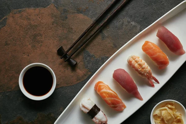 Vista superior de surtido nigiri sushi conjunto, palillos y salsa de soja en plato blanco en la superficie oscura - foto de stock