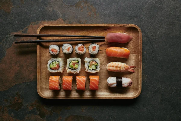 Vista superior do conjunto de sushi arranjado em placa de madeira e pauzinhos na superfície escura — Fotografia de Stock