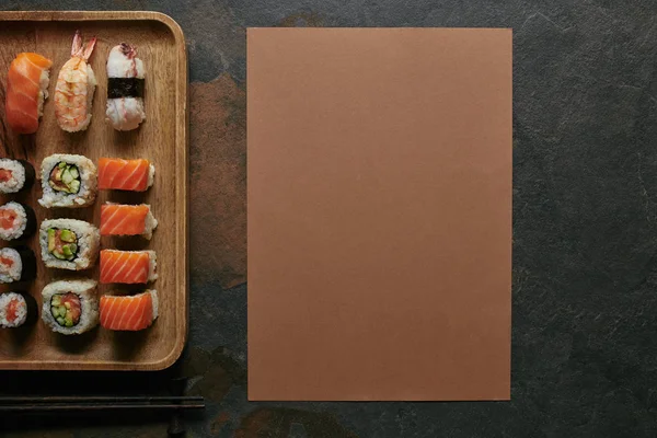 Верхний вид готовых суши на деревянной тарелке и палочки для еды на темной поверхности — стоковое фото