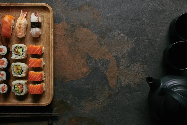 Flat lay com sushi definido em placa de madeira, bule preto e copos em mesa escura — Fotografia de Stock