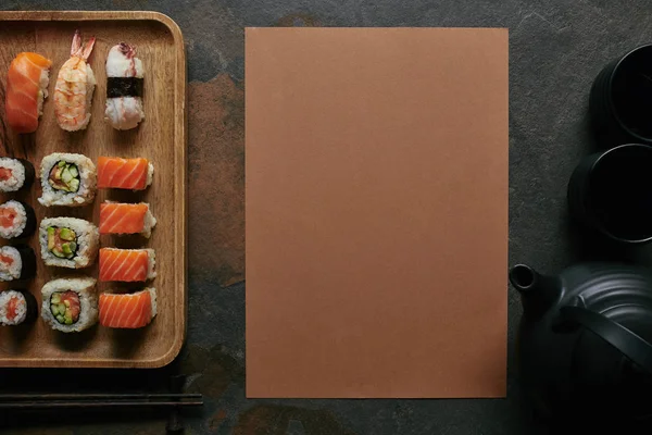 Posa piatta con sushi set su piatto di legno, teiera nera e tazze su tavolo scuro — Foto stock