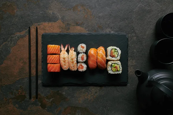 Top view of sushi set on black slate plate, teapot and cups with tea on dark surface — Stock Photo