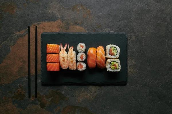 Vue du dessus des sushis disposés sur une plaque d'ardoise noire et des baguettes sur une surface sombre — Photo de stock