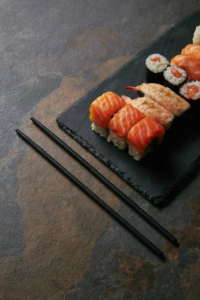 Vista ravvicinata di sushi disposti su piastra di ardesia nera e bacchette su superficie scura — Foto stock