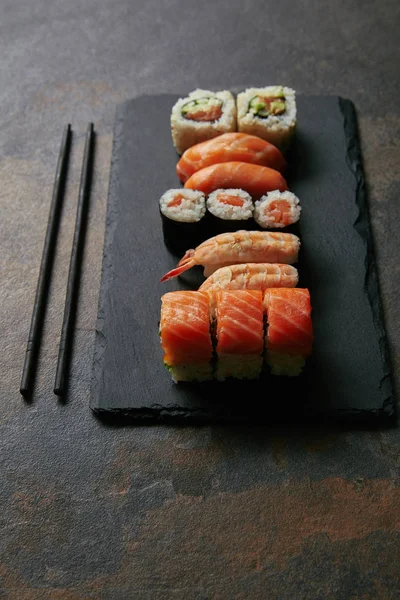 Vista de cerca del conjunto de sushi arreglado en la placa de pizarra negra y palillos en la superficie oscura - foto de stock