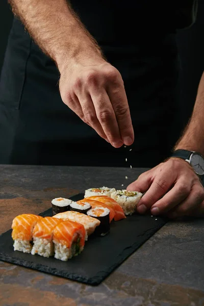 Vista parcial del chef vertiendo semillas de sésamo en sushi arreglado en un plato de pizarra sobre una mesa oscura - foto de stock