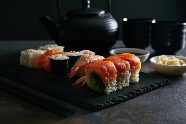 Composición de los alimentos con sushi set, jengibre y salsa de soja en cuencos, tetera y tazas de té en la superficie oscura - foto de stock