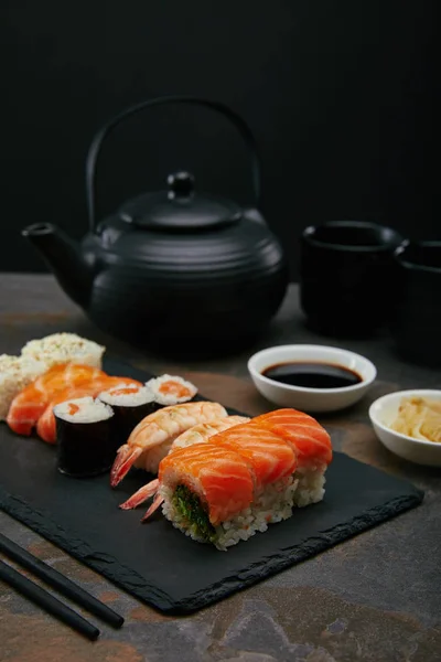Food composition with sushi set, ginger and soya sauce in bowls, teapot and cups with tea on dark surface — Stock Photo