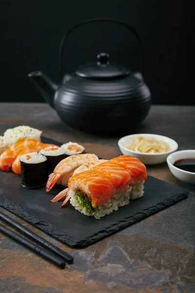 Пищевой состав с набором суши, имбирный и соевый соус в мисках, чайник и чашки с чаем на темной поверхности — стоковое фото
