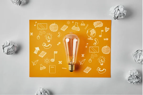 Top view of incandescent lamp on blank yellow paper with business icons surrounded with crumpled papers on white surface — Stock Photo