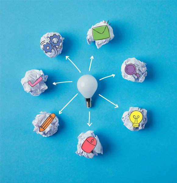 Top view of light bulb surrounded with crumpled papers with business icons on blue surface — Stock Photo