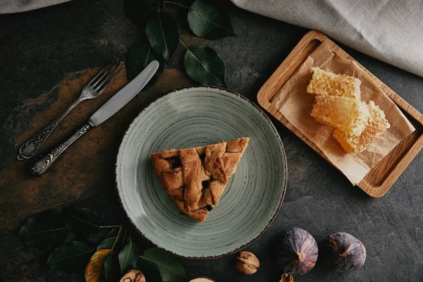 Acostado plano con trozo de pastel en el plato, cera de abejas, higos y cubiertos en la mesa gruesa - foto de stock