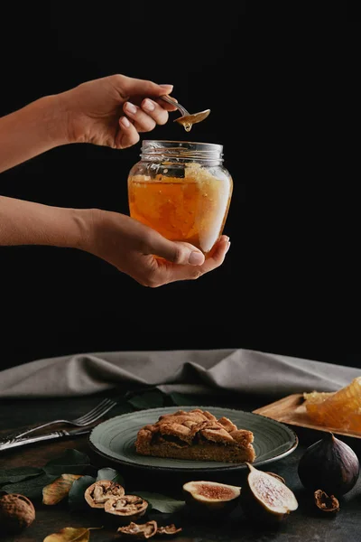 Tiro recortado de mulher segurando frasco de vidro com mel na mesa com pedaço de torta caseira — Fotografia de Stock