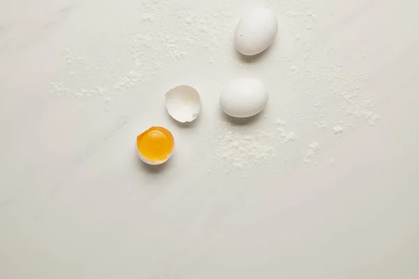Vista superior de huevos de pollo crudos y harina sobre mesa de mármol blanco - foto de stock