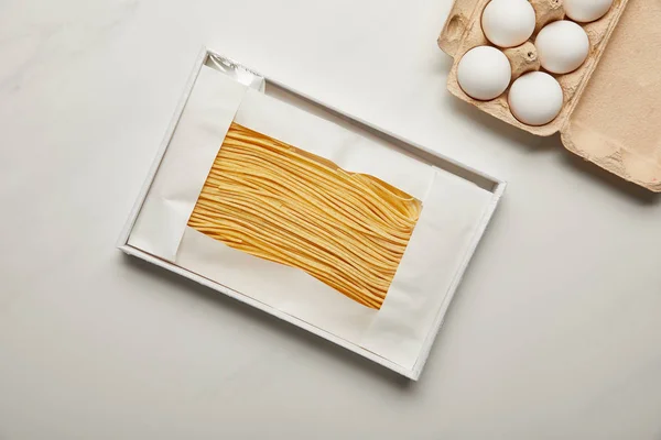 Vista superior de macarrão não cozido e ovos de galinha crus na caixa na superfície de mármore branco — Fotografia de Stock