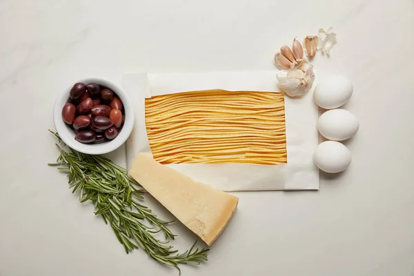 Draufsicht auf verschiedene rohe Makkaroni, Rosmarin, Käse, Knoblauch und rohe Hühnereier Zutaten für Pasta auf weißer Marmoroberfläche — Stockfoto