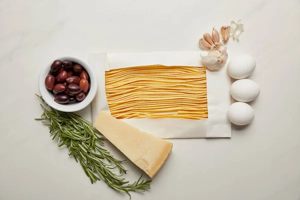 Top view of assorted uncooked macaroni, rosemary, cheese, garlic and raw chicken eggs ingredients for pasta on white marble surface — Stock Photo